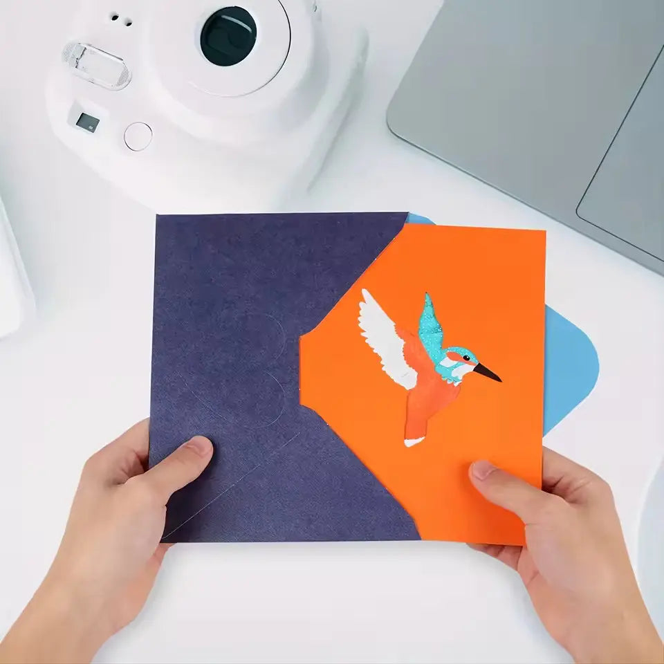 Colibri Memories Album mit buntem Kolibri-Design auf orange-blauem Einband in Händen vor Kamera und Laptop